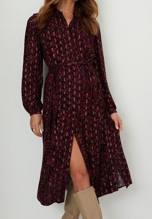 Robe bordeaux à manches longues avec un motif floral, un col et une fente avant, cintrée à la taille, associée à des bottes beiges montantes jusqu'au genou.