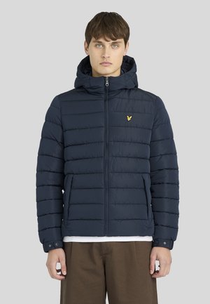 Mann trägt eine marineblaue, gesteppte Jacke mit Kapuze und Reißverschluss, mit einem kleinen gelben Vogel-Logo auf der Brust und braune Hose.