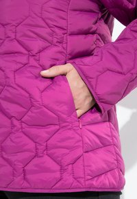 Ruka částečně uvnitř zipované kapsy na rukávu a tělesné části fialového hexagonového prošívaného bundy.