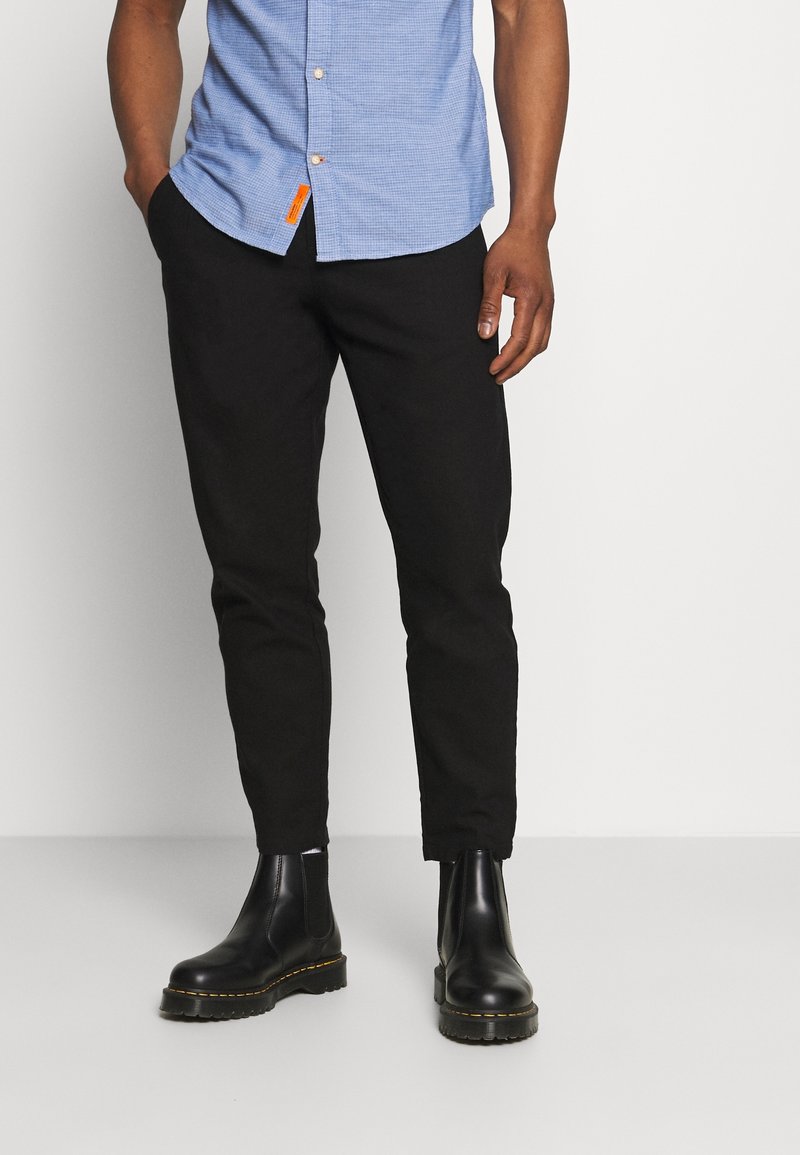 Camicia azzurra a scacchi con bottoni, pantaloni neri affusolati e stivaletti chelsea in pelle nera con cuciture gialle. Dettaglio con etichetta arancione sulla camicia.