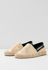Espadrillas metálicas doradas de tipo slip-on con una banda elástica negra que presenta el logo "SUPERDRY". Suela de yute natural con un acabado texturizado.