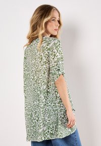 Top leggero verde e bianco con motivi astratti, maniche corte e vestibilità ampia, abbinato a jeans blu a vita alta.