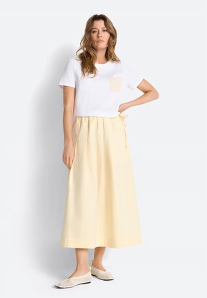 Žena stojaca s rukou v bok, oblečená v bielej krátkoročnej košeli s čipkovanou vreckom, bledozltej midi sukni a béžových sieťovaných balerínach.