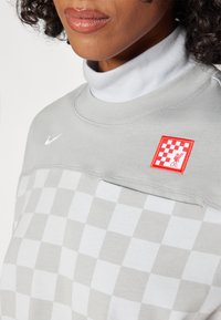 Hanorac gri cu un model în carouri, cu un guler alb înalt dedesubt și un patch roșu brodat LFC cu logo-ul alb Nike în colțul din stânga sus.