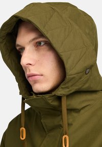 Vadderad jacka med huva i olivgrön, med orange detaljer, dragsko och tryckknappsfunktioner, samt en slät, matt yta.