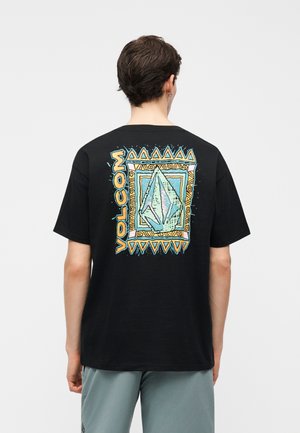 Άτομο που φοράει μαύρο T-shirt με πολύχρωμο γεωμετρικό σχέδιο και το κείμενο «Volcom» στην πλάτη, σε συνδυασμό με ανοιχτό γκρι σορτς.