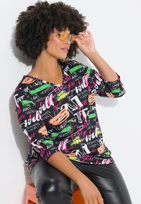 Musta paita, jossa on monivärisiä tekstisuunnitteluja eri fonteilla; kolmannekselle hihoille; sileää kangasta; yhdistetty kiiltäviin mustiin housuihin.