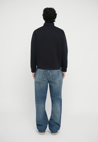 Sweatshirt noir avec col montant, associé à un jean en denim bleu ample. Les jeans ont des poches arrière standard et une texture délavée.