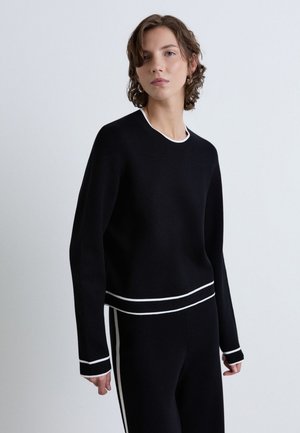 Giovane con capelli corti e ricci indossa una maglia nera a maniche lunghe e pantaloni con bordo bianco, in piedi contro uno sfondo chiaro semplice.