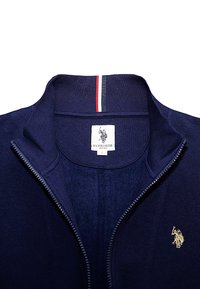 Námornícka bunda na zips s rebrovaným golierom. Na ľavej strane je zlaté logo a na golieri sú červené a biele pruhy.