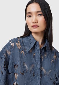Blusa in pizzo blu con motivi floreali, dotata di colletto e chiusura a bottoni. Il design presenta ricami intricati e dettagli a intaglio.