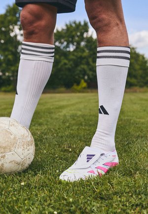 Chaussure de foot blanche avec des accents rose et violet, portée avec des chaussettes blanches montantes, debout sur de l'herbe verte à côté d'un ancien ballon de football.