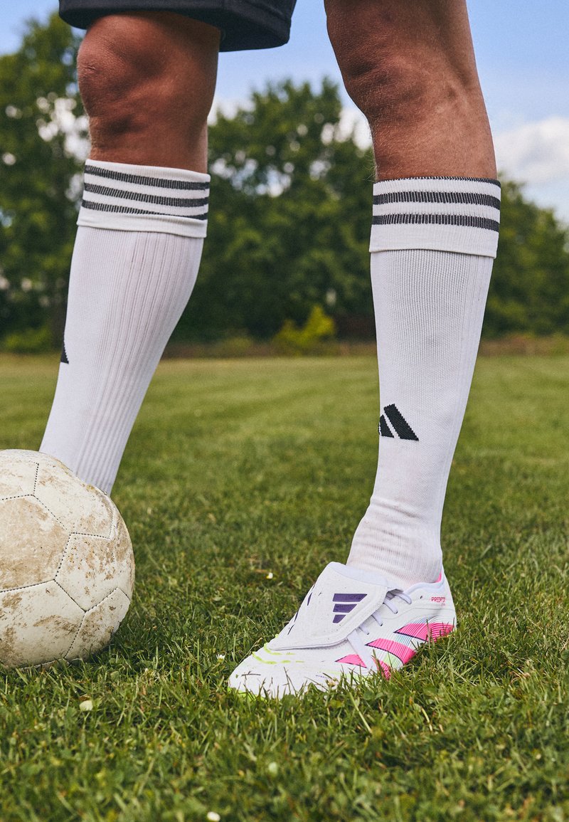 Botín de fútbol blanco con acentos en rosa y púrpura, usado con calcetas blancas hasta la rodilla, apoyado sobre hierba verde junto a un balón de fútbol desgastado.