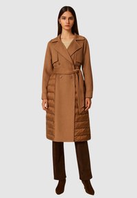 Hnedý, dvojradový trenčkot s vypchávkami v dolnej časti, opaskom a širokými klopami. Model nosí tmavé nohavice a vysoké čižmy.