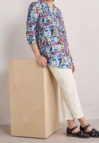 Camicia con motivi colorati e accenti blu, rossi e gialli, con maniche a tre quarti; abbinata a pantaloni crema chiaro e sandali scuri.