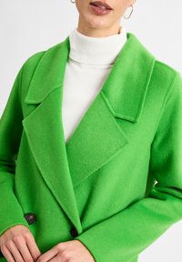 Green wool-blend coat with a wide lapel collar, two buttons, and a textured surface, layered over a white turtleneck.