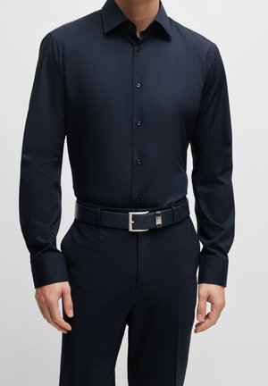 Mies pukeutunut istuvaan tummansiniseen kauluspaitaan, joka on tummansävyisiin housuihin sullottu, vyönään tummansininen vyö hopeisella soljella, seisoo yksinkertaisen taustan edessä.