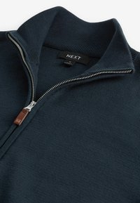 Dunkelblaue Zip-up-Jacke aus strukturiertem Stoff, mit Rippkragen und silbernem Reißverschluss, der ein braunes Lederakzentetikett hat. Markenetikett ist sichtbar im Inneren.