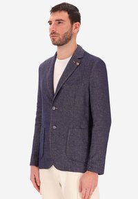 Uomo che indossa un blazer strutturato blu scuro con due bottoni, tasche a toppa e una maglietta bianca a collo rotondo sotto.