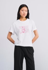 Bílá bavlněná trička s velkým růžovým potiskem s nápisem "PUMA". Otočené krátké rukávy a volný střih, doplněné černými kalhotami.