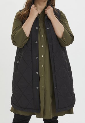 Person iført en sort quiltet vest over en olivengrøn lang skjorte med knapper, der holder vestens krave med begge hænder.