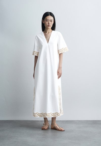 Femme debout portant une robe blanche longue jusqu’aux chevilles avec une bordure brodée dorée sur les manches, l’ourlet et les côtés, associée à des sandales plates couleur beige.