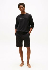 Felpa nera con logo "TOMMY HILFIGER", abbinata a pantaloni corti sportivi neri. Entrambi sono realizzati in morbido tessuto e indossati da un modello.