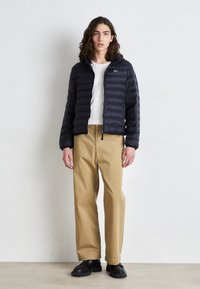 Marinblå puffjacka med horisontell quiltning, dragkedja och huva. Kombinerad med khaki-byxor och svarta skor, står mot en neutral bakgrund.