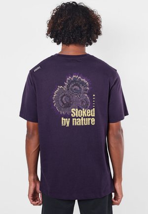Personne aux cheveux bouclés portant un t-shirt violet foncé avec trois motifs de tournesols et le texte "Stoked by nature" au dos.