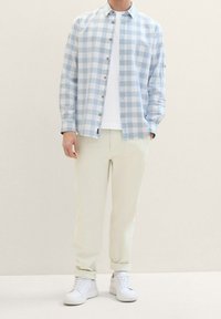 Chemise à manches longues bleu clair à carreaux avec des boutons marron, associée à un pantalon crème et des baskets blanches. Texture lisse et coupe décontractée.