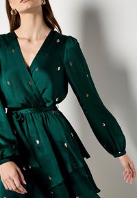 Groene wikkeljurk met lange mouwen, uitgevoerd in een textuurstof met gouden pailletten accents verspreid over de jurk. Tailleband detail.