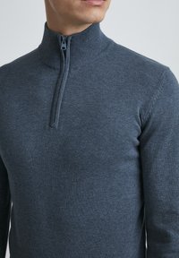 Pullover grigio in maglia con zip, caratterizzato da un colletto alto, dettagli a coste sul collo e una vestibilità aderente. Texture morbida e colore uniforme.