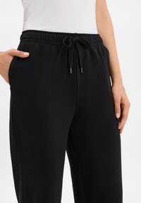 Kobieta ubrana w czarne dresowe spodnie na sznurku, z rękami w pobliżu kieszeni, oraz w białą bezrękawnikową bluzkę na jednolitym tle.