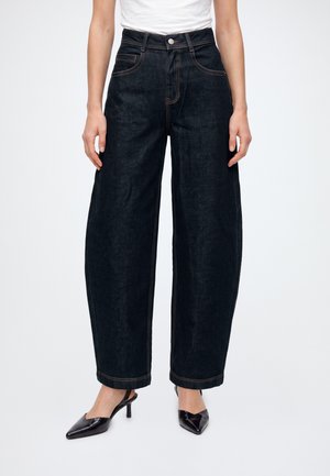 Pessoa a usar jeans escuros de cintura alta e pernas largas, e saltos slingback pretos com bico fino, em pé contra um fundo liso.