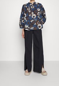 Blusa con stampa floreale in blu, marrone e avorio con collo alto, abbinata a pantaloni neri larghi con aperture laterali e sandali platform beige.