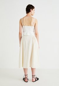 Hvit smocket crop-top med blonde detaljer og volangkant, kombinert med en kremfarget, midilengde skjørt. Svarte sandaler med stropper fullfører antrekket.