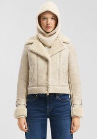 Vrouw in een beige schapenvachtjack met een brede kraag en manchet, een bijpassende gebreide capuchon en donkerblauwe jeans.