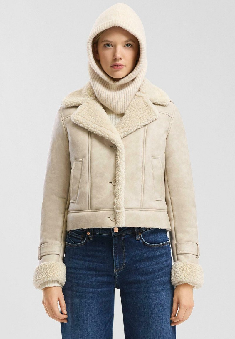 Vrouw in een beige schapenvachtjack met een brede kraag en manchet, een bijpassende gebreide capuchon en donkerblauwe jeans.