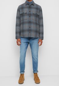 Giacca a camicia grigia a quadri con tasche sul petto, chiusura con bottoni sul davanti e una texture morbida, abbinata a jeans azzurri chiari e stivali color miele.