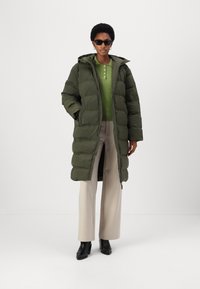 Olive green puffer coat with a hood, zip closure, and quilted texture. Paired with a light green top and beige wide-leg trousers.