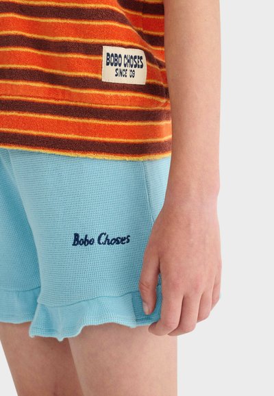 Enfant portant un T-shirt rayé orange et marron et un short bleu clair texturé avec la marque "Bobo Choses" sur les deux vêtements.