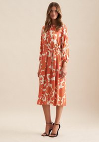 Blumenkleid in Koralle und Creme, mit langen Ärmeln, elastischem Bund und fließendem Stoff. Kombiniert mit schwarzen High-Heel-Sandalen.