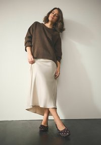 Brązowy, oversize’owy sweter, kremowa satynowa spódnica midi z asymetrycznym dołem oraz czarne baleriny w białe groszki. Gładkie tekstury i delikatne kolory.