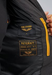 Zwarte jasvoering met een oranje rits, stoffen label met de tekst "PME LEGEND" en "YETCRAFT", en subtiele stikdetails.