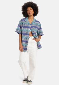 Camisa de mangas cortas en azul con patrones geométricos, combinada con pantalones anchos blancos y zapatillas negras. Material de algodón, corte relajado.