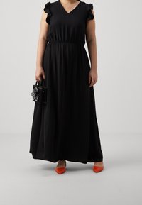 Kvinna klädd i en svart maxiklänning med volangärmar, som håller en liten svart handväska med blomdetalj och har på sig orange spetsiga skor.