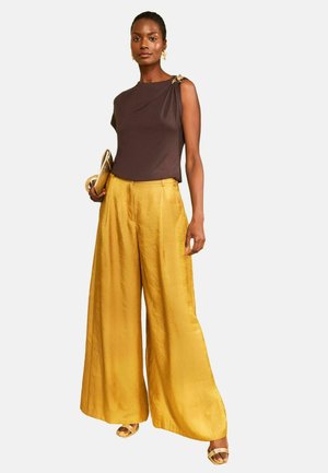 Žena nosiaca tmavohnedý tielkový top so zlatou brošňou, široké lesklé zlaté nohavice, zlaté opätky, drží zlaté psaníčko v ľavej ruke.