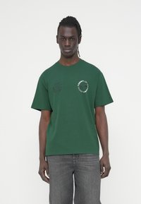 Πράσινο βαμβακερό μπλουζάκι με στρογγυλή λαιμόκοψη, κοντά μανίκια και τυπωμένη κυκλική λεπτομέρεια με λογότυπο στο μπροστινό μέρος, συνδυασμένο με σκούρα γκρι τζιν.