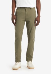 Chino khaki con vestibilità dritta, realizzati in cotone, dotati di tasche laterali e una texture liscia, abbinati a sneaker bianche.