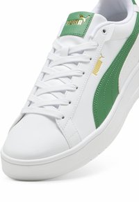 Sneaker bianca con texture in pelle liscia, caratterizzata da dettagli verdi, un logo dorato sulla linguetta e una suola bianca imbottita.